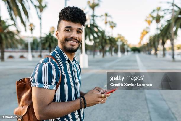 smiling young man holding smartphone standing outdoors - latin american and hispanic ethnicity on phone stock pictures, royalty-free photos & images