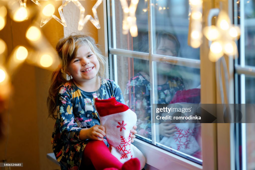 Little preschool girl holding cup Santa Claus boot with gift called Nikolausstiefel in German. Happy child wait on holiday by window with Christmas lights in winter. Cozy family celebration of xmas.