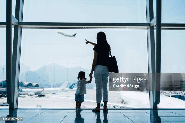 rear view of young asian mother holding hands of cute little daughter looking at airplane through window at the airport while waiting for departure. family travel and vacation concept - kid in airport stock-fotos und bilder