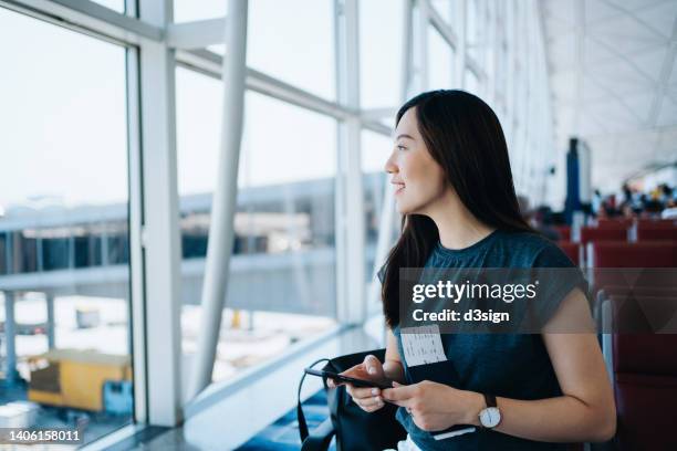 smiling young asian woman holding flight ticket and passport on hand, looking through the window while using smartphone and waiting for her flight in airport lounge. travel and vacation concept. business person on business trip - funkenflug stock-fotos und bilder