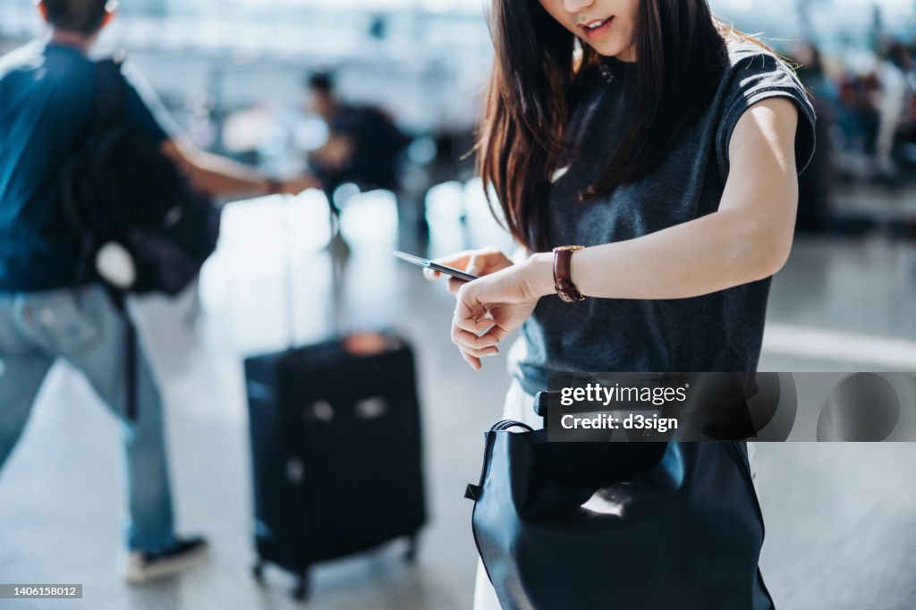 Cropped shot of young Asian woman with suitcase and passport, checking time on wristwatch in the airport concourse. Travel and vacation concept. Business person on business trip