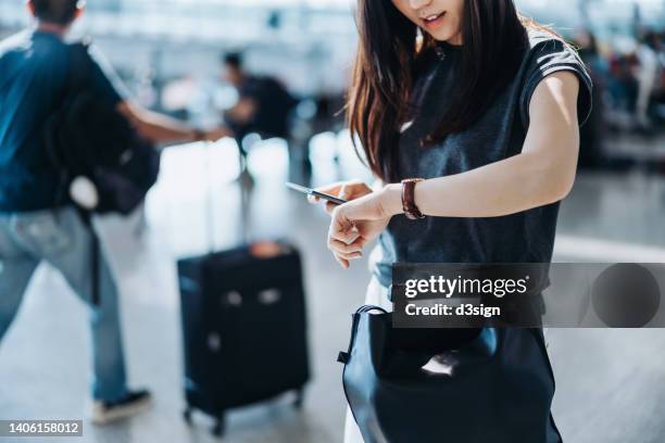 cropped shot of young asian woman with suitcase and passport, checking time on wristwatch in the airport concourse. travel and vacation concept. business person on business trip - cancellation stock pictures, royalty-free photos & images