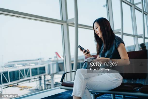 young asian businesswoman holding flight ticket and passport on hand, using smartphone while waiting for her flight in airport lounge. travel and vacation concept. business person on business trip - passenger stock-fotos und bilder