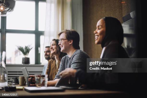 cheerful young businessman sitting by male and female colleagues while discussing in meeting room - ftm stock-fotos und bilder