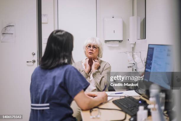 senior woman explaining throat problem to otolaryngologist sitting at desk in medical clinic - médico de cabecera fotografías e imágenes de stock