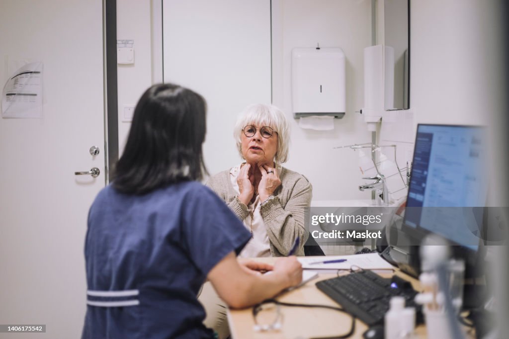 Senior woman explaining throat problem to otolaryngologist sitting at desk in medical clinic