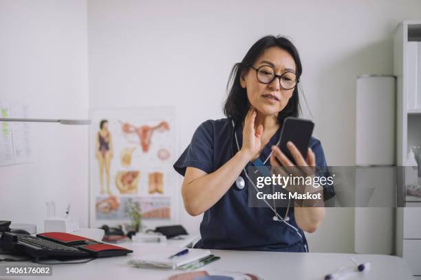 female otolaryngologist doing online consultation through mobile phone while sitting at desk in medical clinic - telemedicine stock pictures, royalty-free photos & images