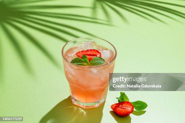 drinking glass of strawberry cocktail with mint leaves and iced cubes on bright green background, tropical tree shadow. homemade ready-to-eat lemonade. vitamin delicious refreshing drink, horizontal, copy space - cocktail recipe stock pictures, royalty-free photos & images