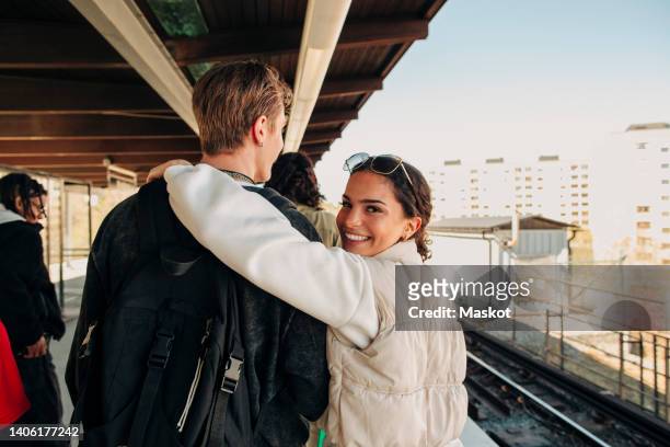 portrait of smiling woman with arm around friend walking on railroad station - man looking back stock pictures, royalty-free photos & images