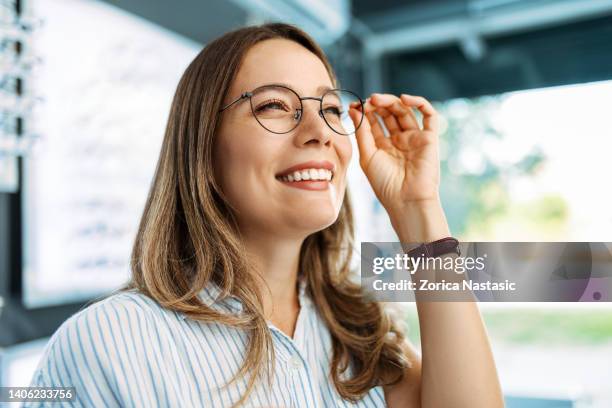 une jeune femme souriante choisit des lunettes dans un magasin d’optique - lunettes de vue photos et images de collection