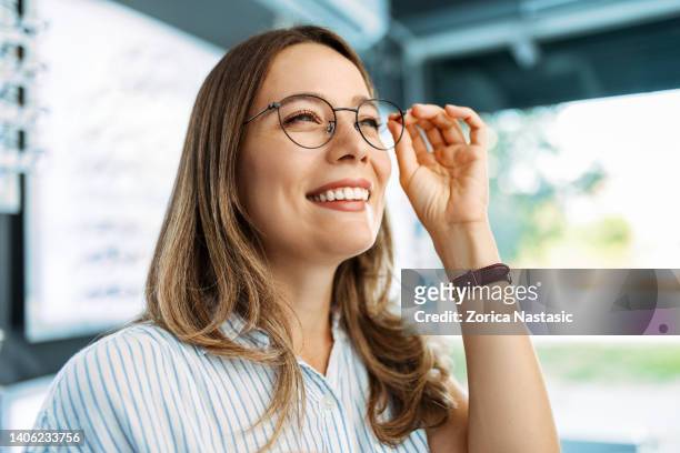 smiling young woman choosing eyeglasses in optical store - optical equipment stock pictures, royalty-free photos & images
