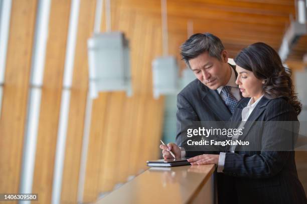 mature caucasian businesswoman and chinese asian businessman standing on office balcony taking notes - government contract stock pictures, royalty-free photos & images