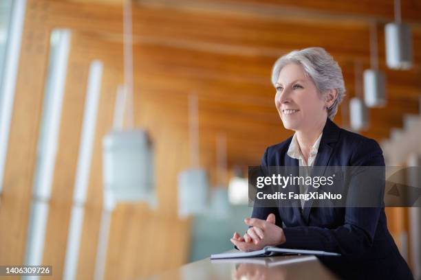 portrait of smiling happy senior caucasian businesswoman leaning on railing at office balcony with hands clasped - politician stock pictures, royalty-free photos & images