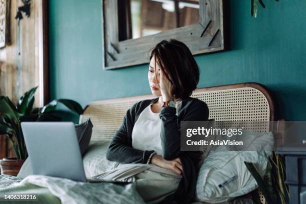 worried young asian woman with her hand on head, using laptop computer on the bed at home, looking concerned and stressed out - dolours price stock pictures, royalty-free photos & images