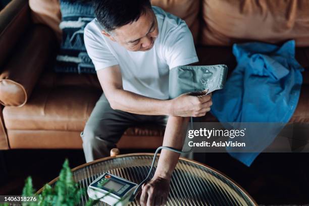 high angle shot of senior asian man checking his blood pressure at home on his arm with a blood pressure monitor. elderly and healthcare concept - high blood pressure stockfoto's en -beelden