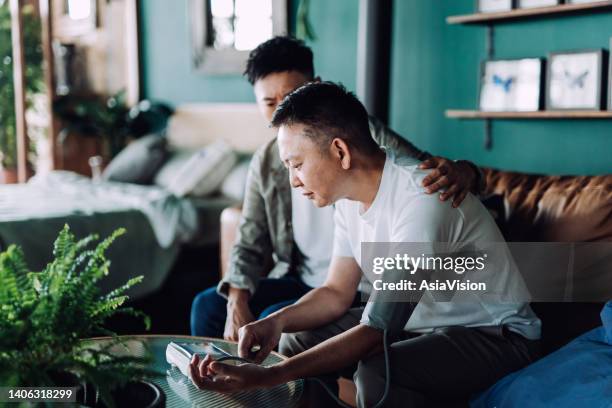 senior asian man checking blood pressure with his son at home. looking worried. son caring for his retired father, putting hand on his shoulder, comforting and consoling his father. family love, care, elderly and healthcare concept - hand på axel bildbanksfoton och bilder