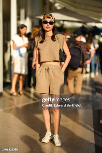 Model wears a black with gold and brown print pattern silk scarf as a hat, black sunglasses, a beige sleeveless / cropped t-shirt, matching sport...