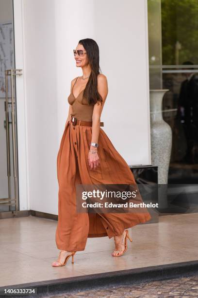 Guest wears beige sunglasses, a large gold and silver chain necklace, a brown ribbed V-neck / tank-top, a brown shiny leather large belt, a brown...