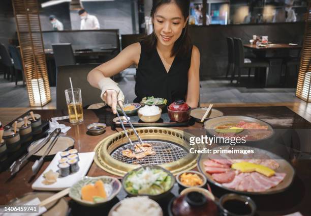 cropped shot young asian woman eating traditional japanese style beef barbecue "yakiniku" in a restaurant and putting raw beef slices over the charcoal grill stove - hot pots stock pictures, royalty-free photos & images