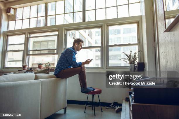 wide shot of young man using smart phone while sitting in living room - 2015 18 stock pictures, royalty-free photos & images