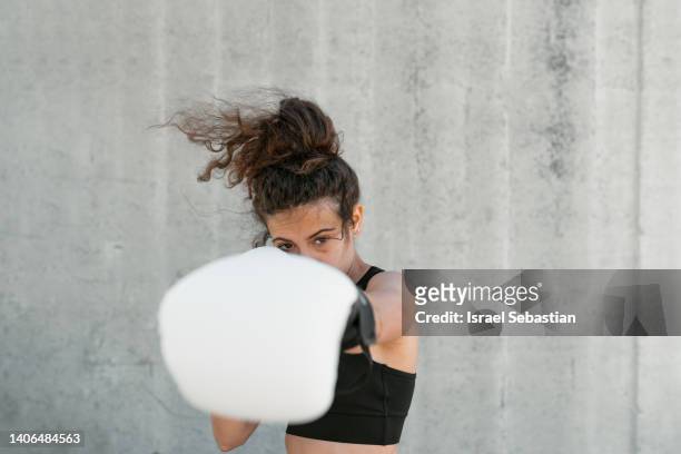 woman boxer with with boxing gloves standing in fighting stance and looking at camera during training against gray wall. - boxing glove stockfoto's en -beelden