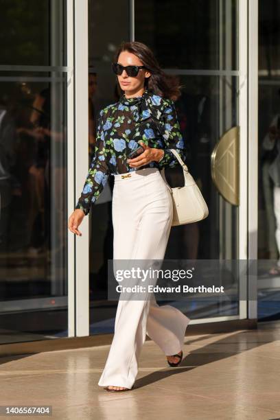 Guest wears black sunglasses, a black tulle with green and blue flower print pattern high neck / long sleeves blouse, a white latte varnished leather...