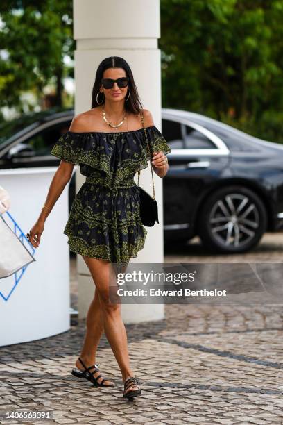 Guest wears black sunglasses, gold earrings, a gold necklace, a black with green print pattern shoulder-off / short sleeves / short jumpsuit, a black...
