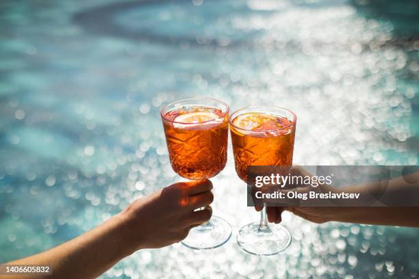 two hands toasting with glasses spritz cocktails on background of swimming pool. cheers - holiday cocktail party stockfoto's en -beelden