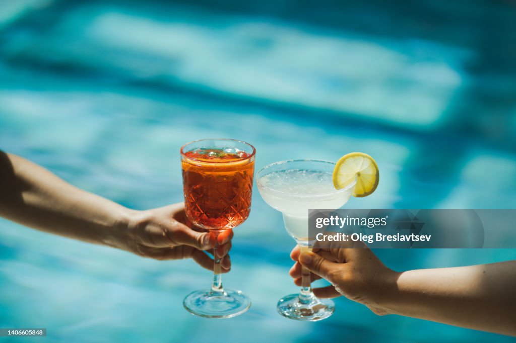 Two hands toasting with glasses Spritz and Margarita cocktails on background of swimming pool. Cheers