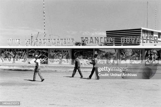 Men walkign down the road, the lettering in the background reading Live, peacebringer Francois Duvalier, Haiti, 1966.