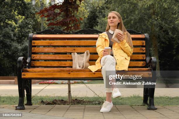 a beautiful young woman or girl holds a disposable mug of coffee in her hands, drinks it, eats a delicious vegetarian or vegan shawarma doner kebab while sitting on a park bench. the concept of fast food and unhealthy food, snacking. ethical consumerism. - park bench stock pictures, royalty-free photos & images