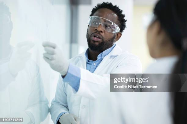 efficiency over thoroughness in laboratory testing decision. male african american scientist leading discussion of experiment with colleagues at white board in laboratory. - microbiologist fotografías e imágenes de stock