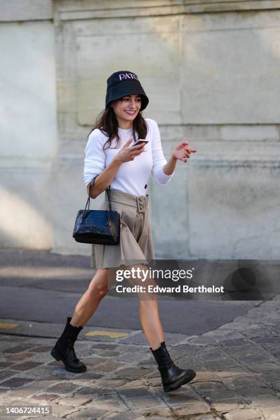 Guest wears a black with embroidered white inscription bob hat from Patou, a white ribbed long sleeves t-shirt, a beige pleated / belted / buttoned...