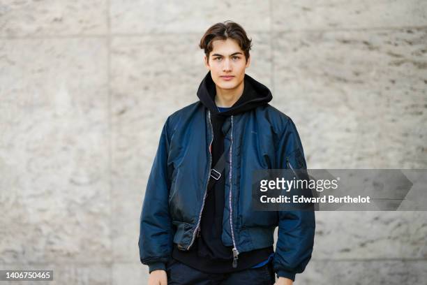 Guest wears a navy blue t-shirt, a black hoodie sweater, a navy blue nylon bomber coat, black large pants, a black crossbody bag, outside the Paco...