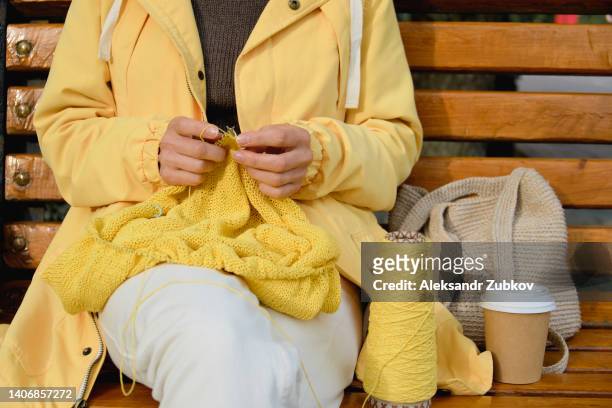 a young woman is knitting a woolen jacket or clothes with knitting needles, sitting on a bench in a public park. the girl holds a bright yarn in her hands and drinks coffee from a disposable cup. hobby concept, freelance creative work. do what you love. - yellow hat stock pictures, royalty-free photos & images