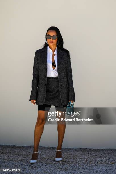 Amina Muaddi wears black sunglasses, silver and rhinestones earrings, a white shirt, a black lace bra underwear, a dark gray blazer jacket from Dior,...