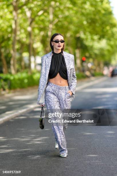Mary Leest wears black sunglasses, a black halter-neck / wrap cropped top, a silver sequined blazer jacket, matching silver sequined suit pants, a...