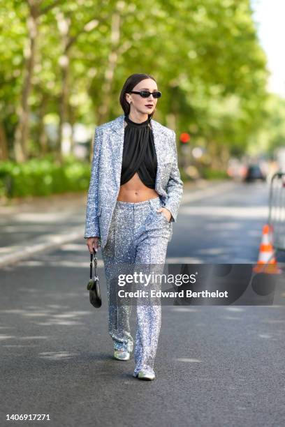 Mary Leest wears black sunglasses, a black halter-neck / wrap cropped top, a silver sequined blazer jacket, matching silver sequined suit pants, a...