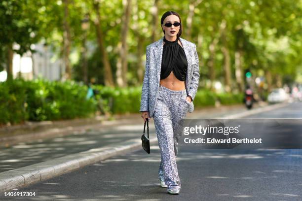 Mary Leest wears black sunglasses, a black halter-neck / wrap cropped top, a silver sequined blazer jacket, matching silver sequined suit pants, a...