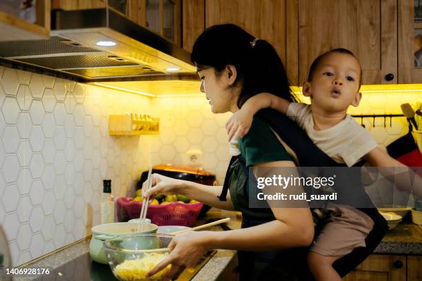 woman cooking in kitchen, baby strapped to body in sling - mãe dona de casa imagens e fotografias de stock