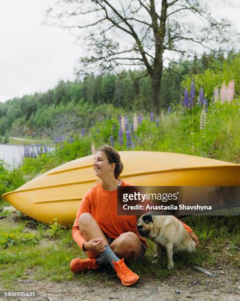 happy female sitting by the yellow canoe and lupine flowers by the lake petting a dog - emotion meer stockfoto's en -beelden