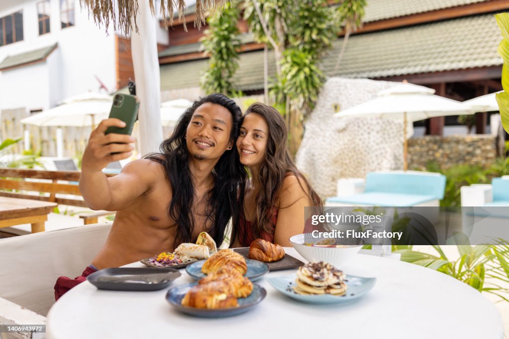Multiracial couple taking selfie, during breakfast at the beach restaurant