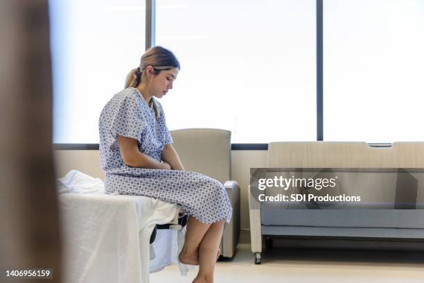 anxious, sad, young woman wearing hospital gown looks down - patient in bed stockfoto's en -beelden