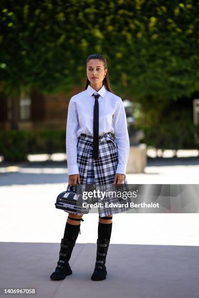 Sarah Lysander wears gold pendant earrings, a white shirt, a black tie, a black and white checkered print pattern slit knees skirt from Dior, a black...