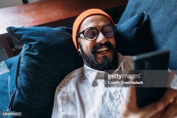a happy handsome man with glasses and an orange cap on his head holding his mobile phone while lying on the sofa - mobile phone at home bildbanksfoton och bilder