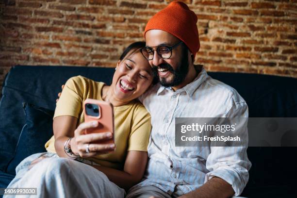 a happy mixed-race couple watching something on the mobile phone while enjoying spending time together at home and building their relationship - sikhisme stockfoto's en -beelden