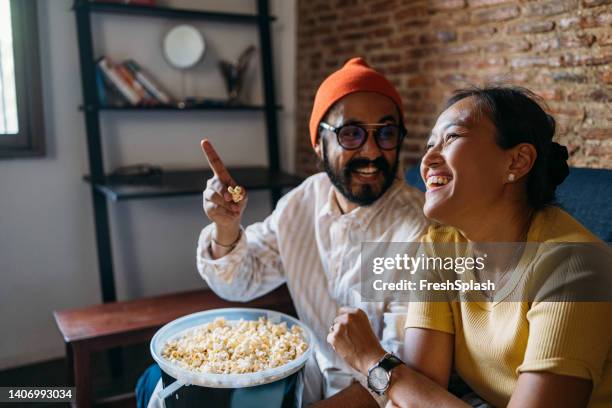 a happy mixed-race couple enjoying eating popcorn while relaxing at home - asian married stock pictures, royalty-free photos & images