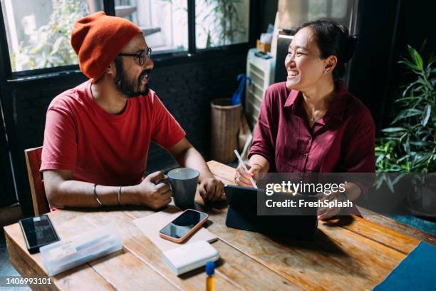 un couple métis souriant assis à table et travaillant à domicile à l’aide de leur tablette - café rouge photos et images de collection