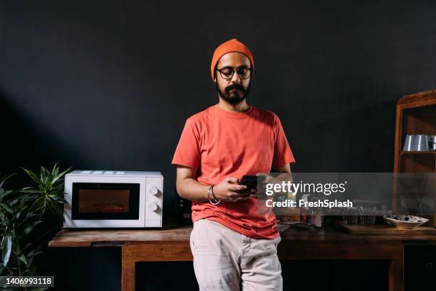 a handsome serious businessman standing in the kitchen and texting on his mobile phone while waiting his lunch in the microwave to be heated - white hat fashion item stockfoto's en -beelden