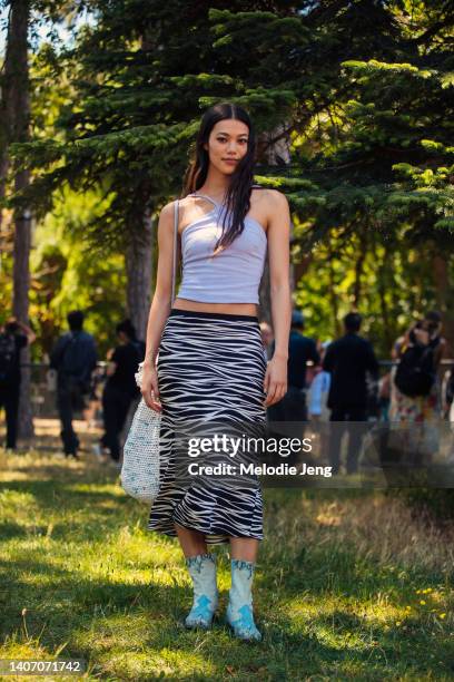 Model Jade Nguyen wears a light purple criss-cross cropped tank top, white horizontal zebra print long midi skirt, blue cowboy boots, and a white big...